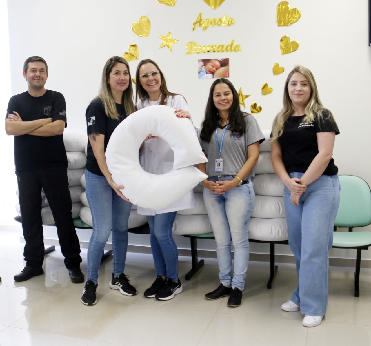um homem de roupas pretas e quatro mulheres. Duas delas seguram uma almofada de amamentação branca
