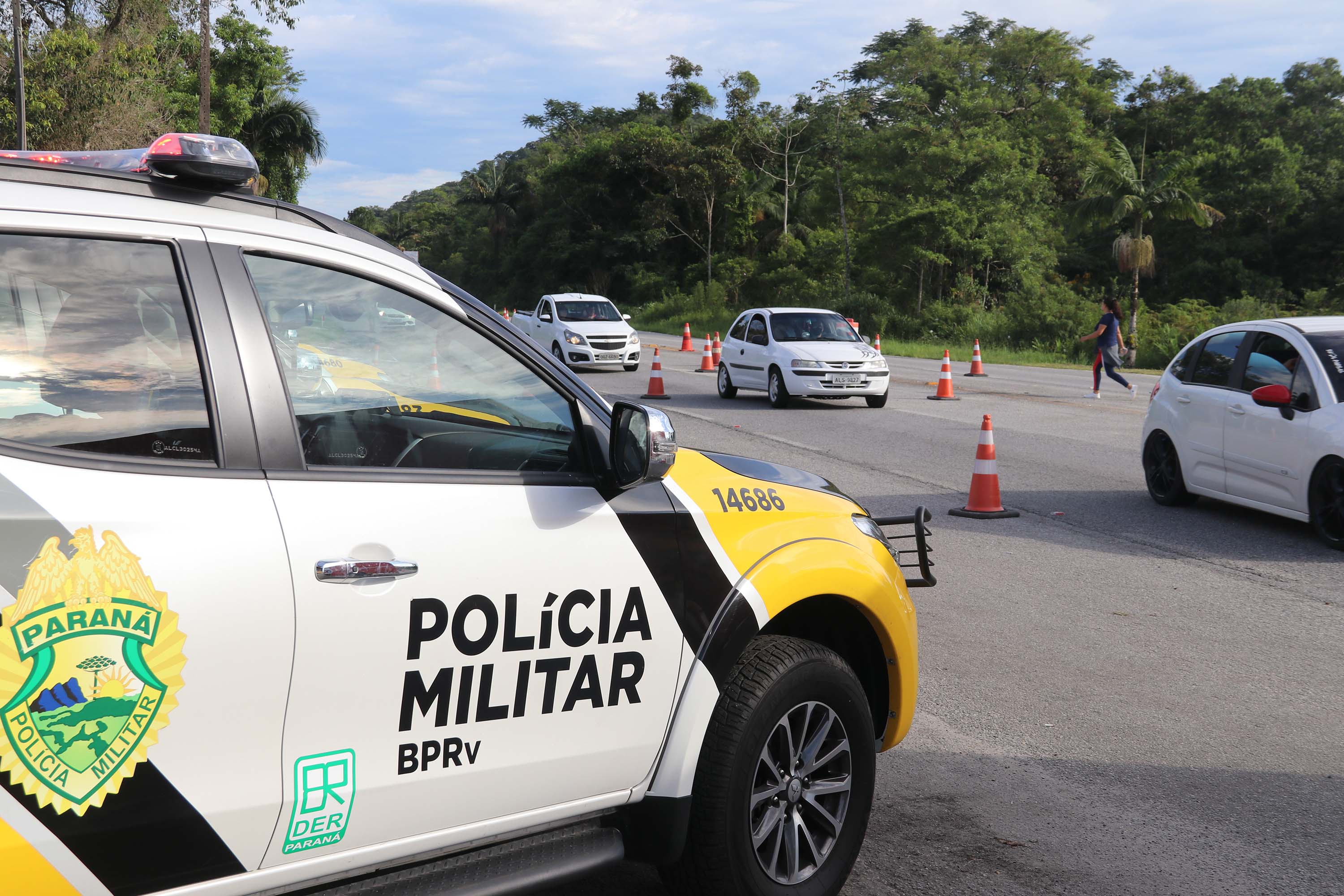 lateral da viatura da PMPR parada em frente á rodovia onde passam carros