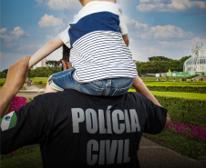 Homem usando camiseta da Polícia Civil carrega uma criança em seus ombros. Ao fundo, a paisagem do Jardim Botânico de Curitiba