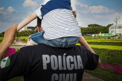 Homem usando camiseta da Polícia Civil carrega uma criança em seus ombros. Ao fundo, a paisagem do Jardim Botânico de Curitiba