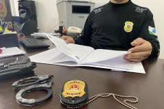 Policial civil segura manual de atendimento. À sua frente está uma algema e um distintivo da Polícia Civil