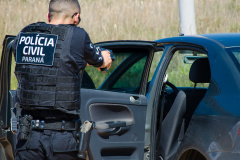 agente da polícia civil de costas para a foto. Ele está em frente à lateral de uma viatura de portas abertas