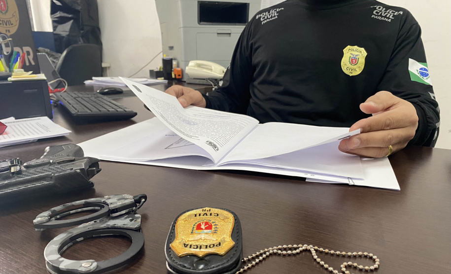 Policial civil segura manual de atendimento. À sua frente está uma algema e um distintivo da Polícia Civil