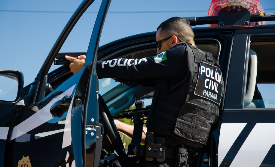 policial civil aparece de lado na imagem. Ele está fardado, ao lado de uma viatura