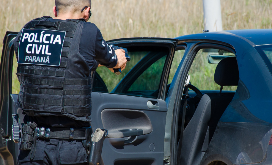 agente da polícia civil de costas para a foto. Ele está em frente à lateral de uma viatura de portas abertas